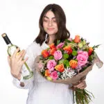 תמונה של דוגמנית מחזיקה ביד אחת זר פרחים צבעטני אהבה בשדות וביד השניה מחזיקה בקבוק יין לבן
